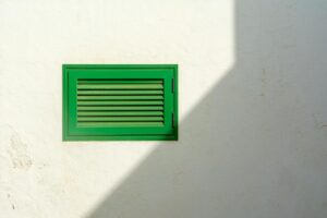 Green vent on a white wall with sharp sunlight casting a shadow, creating a minimalist urban scene.