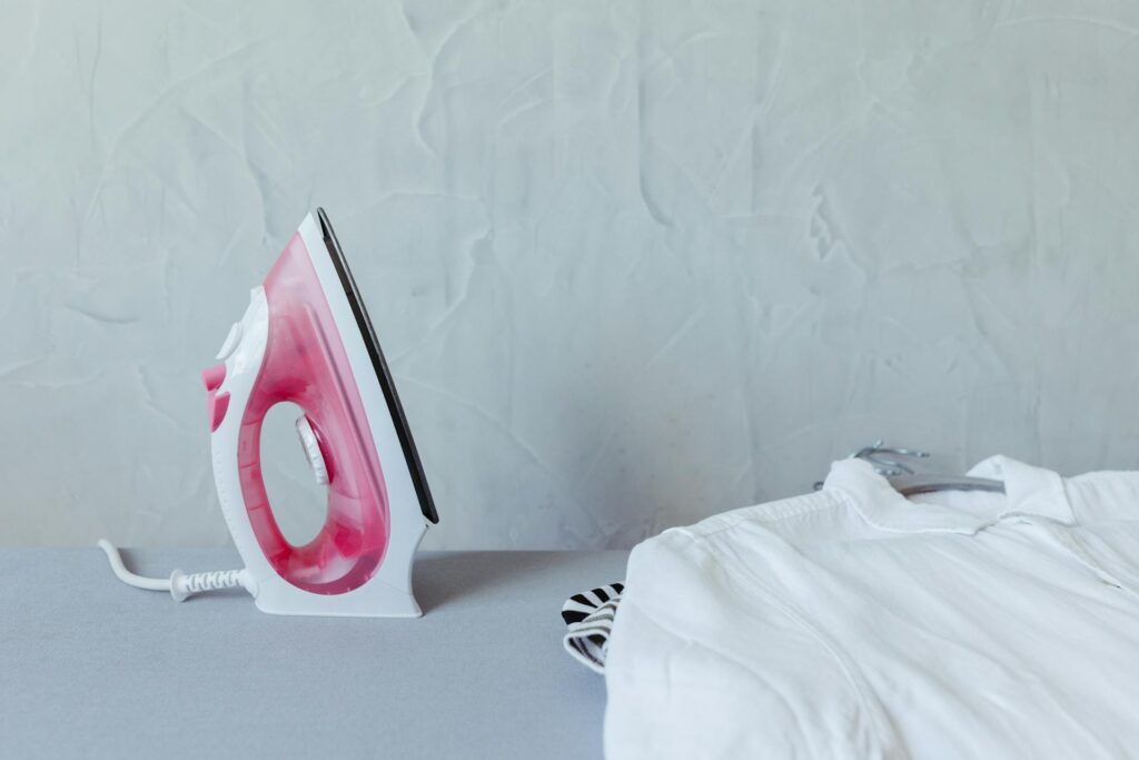 A vibrant pink iron next to a neatly folded white shirt on an ironing board against a textured wall.