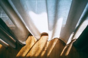 How to Wash Curtains at Home Warm sunlight beams cast patterns through translucent curtains on a wooden floor.