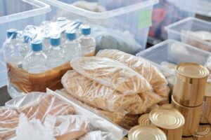 How to Set Up a Basic Emergency Supply Kit Plastic-wrapped food items and bottled water organized in storage containers and tin cans.